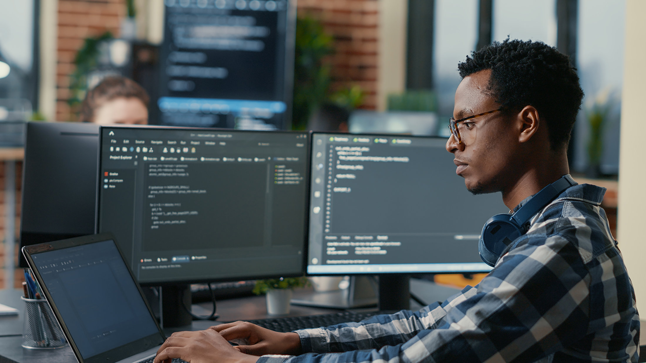 Person wearing a flannel and glasses types code on to a laptop connected to two other monitors in an office setting.