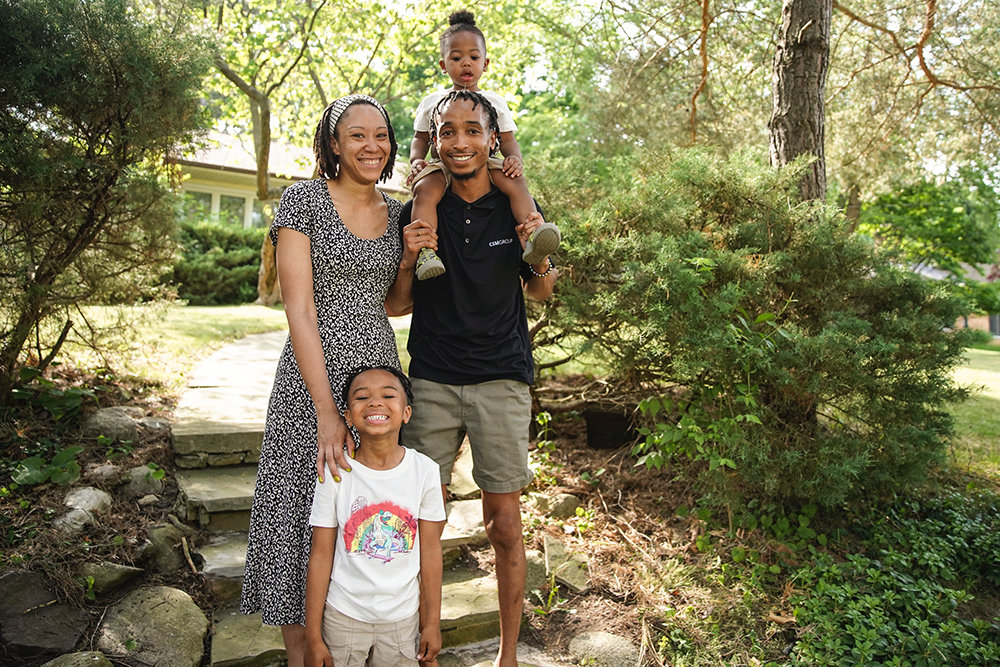 Two adults standing outside on steps with one smiling child standing next to them and another child sitting on the man's shoulders